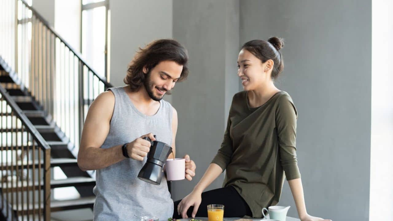 A man smiling while handing his partner a coffee in the kitchen during a relaxed morning.