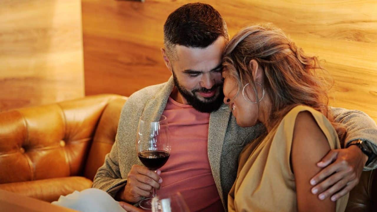 Couple sitting close on a couch, smiling and drinking wine in a quiet, cozy moment.