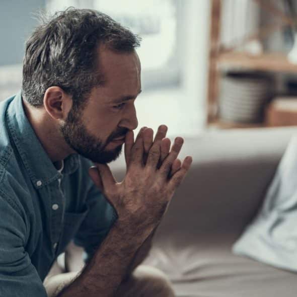 A pensive man with a beard sits on a couch, looking down with his hands clasped.
