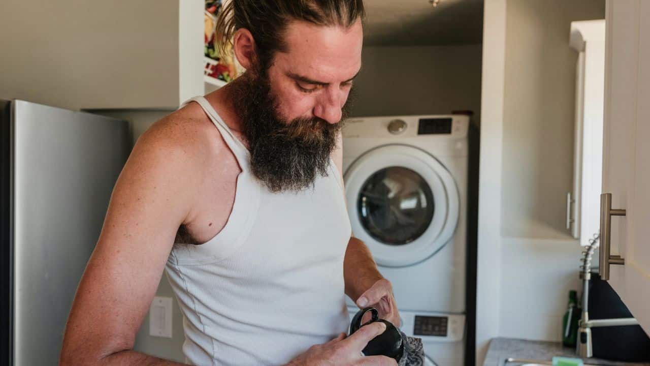 A man with a long beard and hair tied back is washing a black mug in a kitchen with a washing machine in the background.