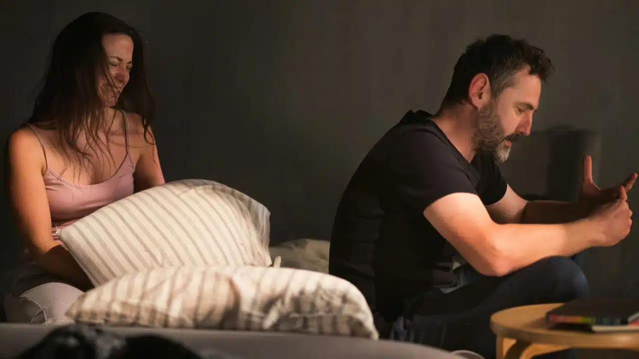 A man and woman sit apart on a couch, the man looking down and the woman smiling with a pillow in front of her.