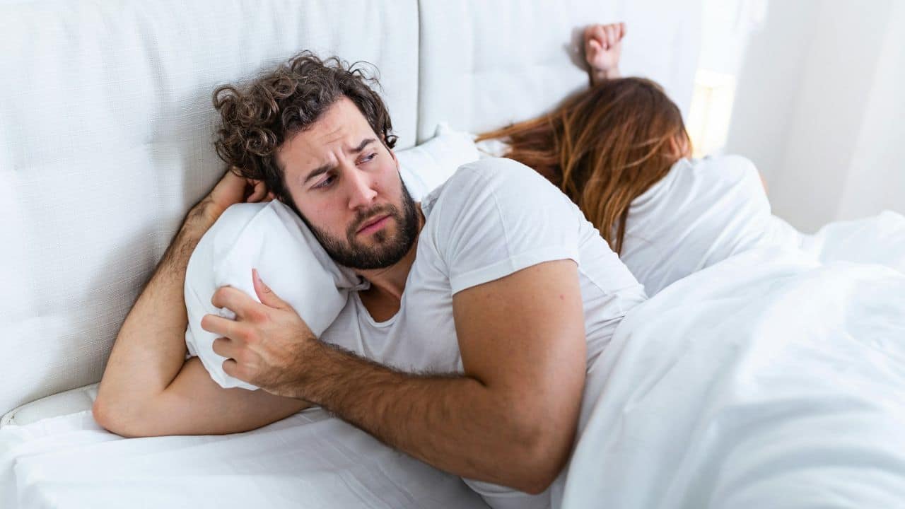 A man with a beard lies in bed looking unhappy, while a woman lies with her back to him.