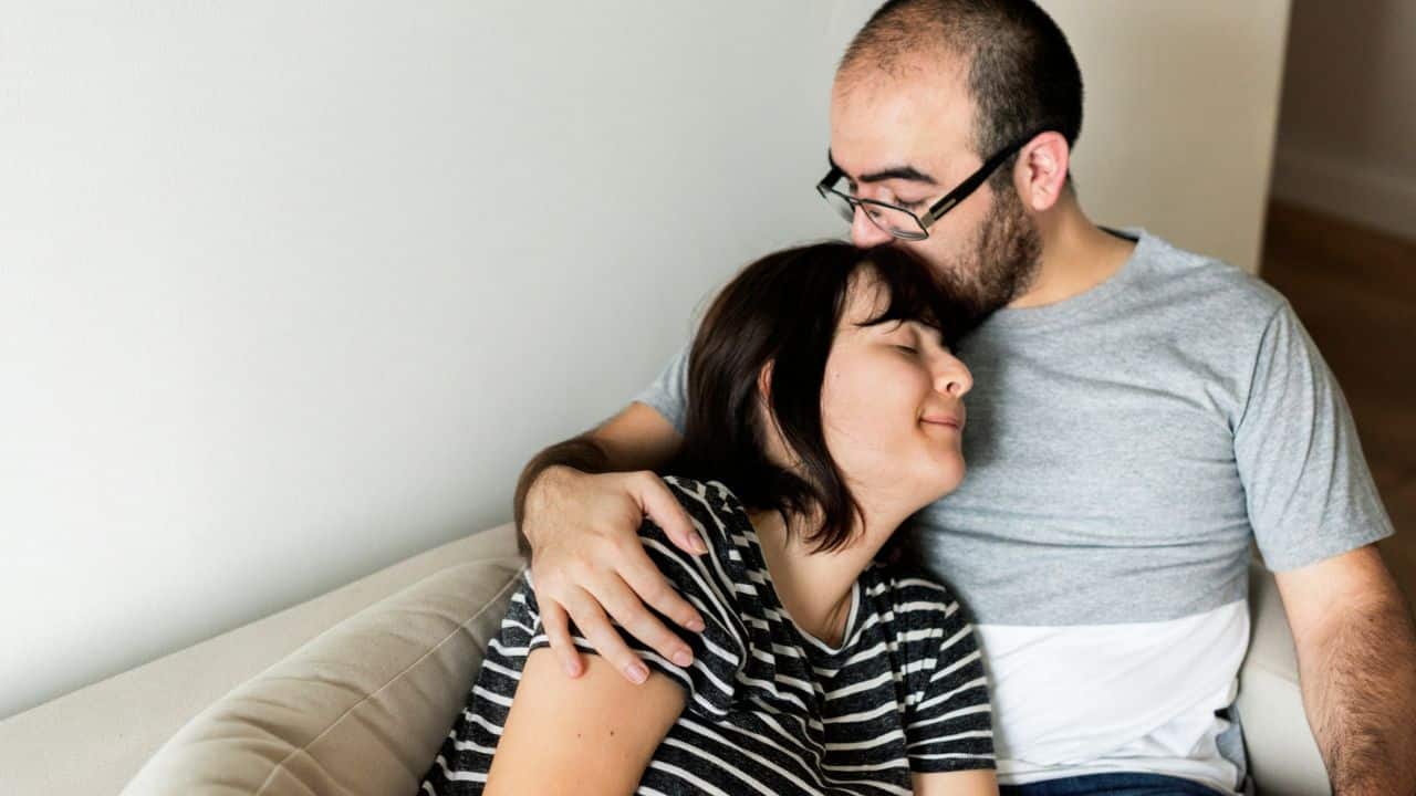 A man with glasses embraces a woman, who leans into his shoulder with her eyes closed.