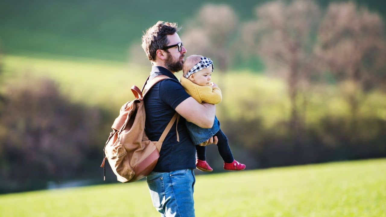 A man with a backpack carries a young child in his arms across a green field.