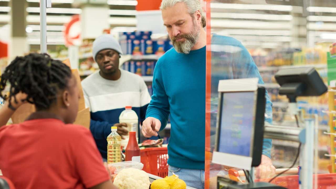A man with a beard and gray hair stands at a checkout counter, looking at items, with a cashier and another customer behind him.