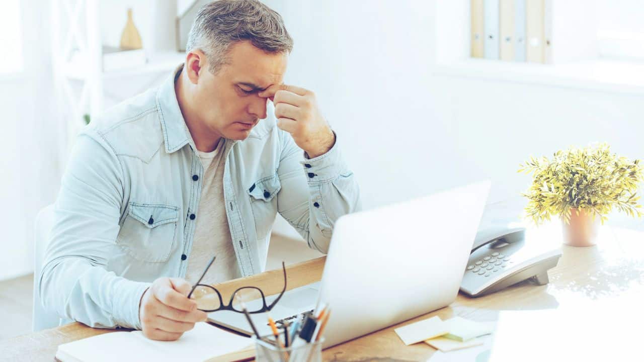 A man in a denim shirt sits at a desk with a laptop and phone, holding glasses and rubbing his forehead.