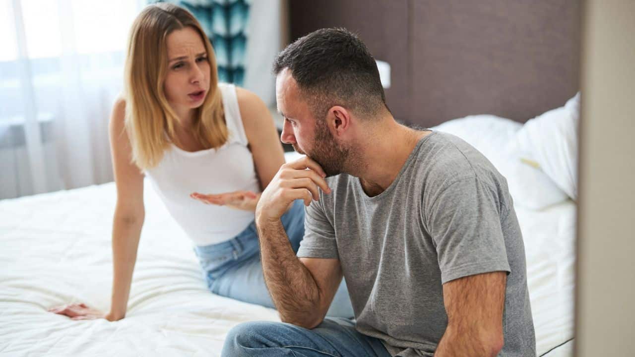 A woman gestures and looks upset at a man who sits with his back to her, looking away thoughtfully.