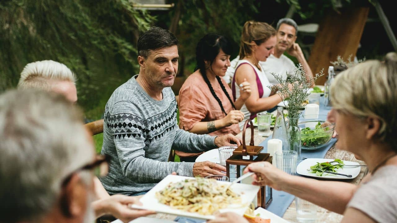 A diverse group of adults is gathered around an outdoor dining table, with one person handing a plate of food to another.
