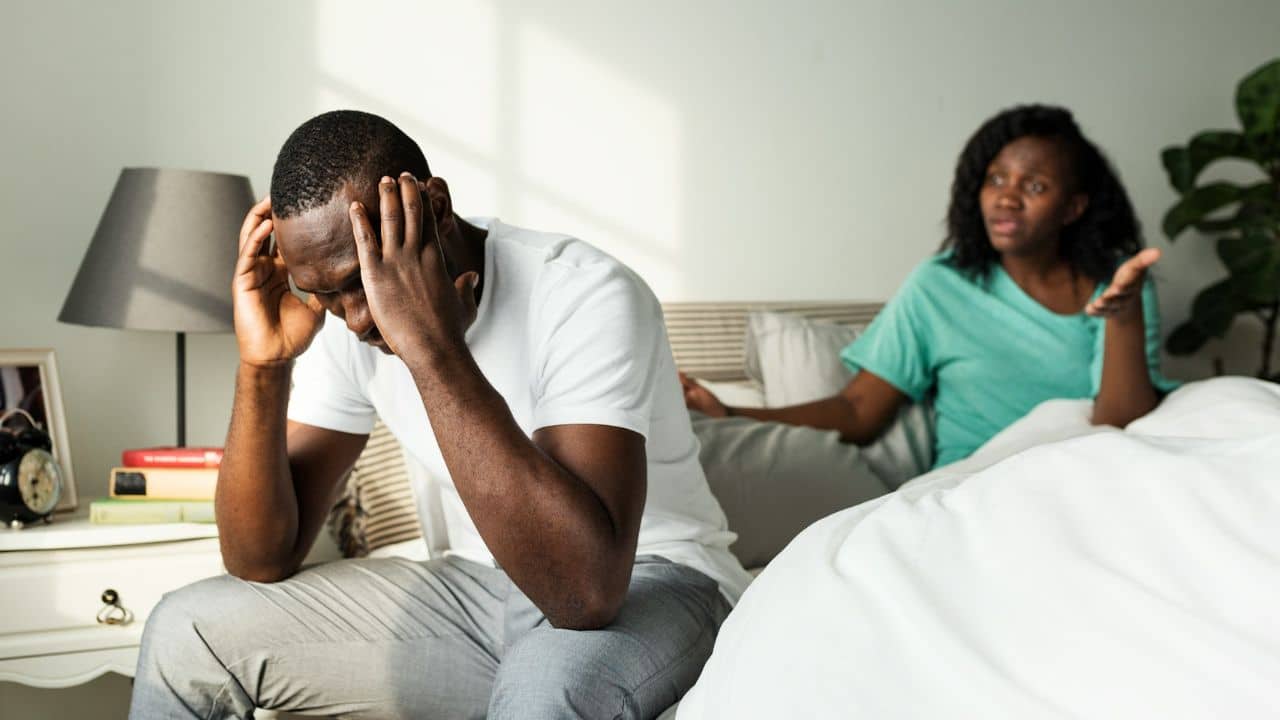 A distressed man sits on the edge of a bed with his head in his hands, while a woman gestures and talks from the bed behind him.