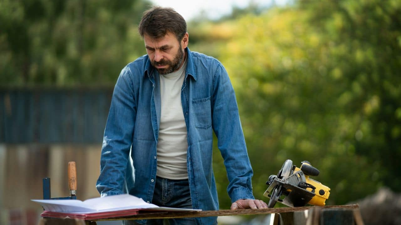 A bearded man in a denim shirt looks down at blueprints on a table with a circular saw nearby outdoors.