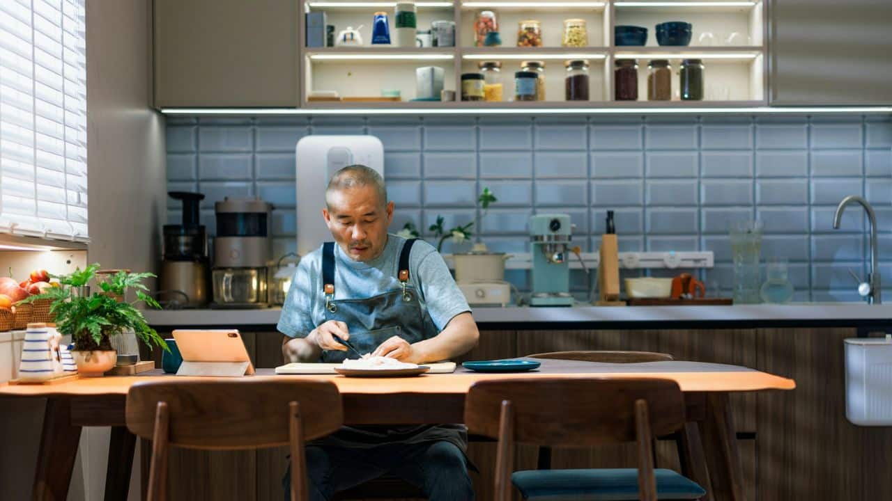 A man in an apron is seated at a kitchen table, looking at a tablet and cutting something on a plate.