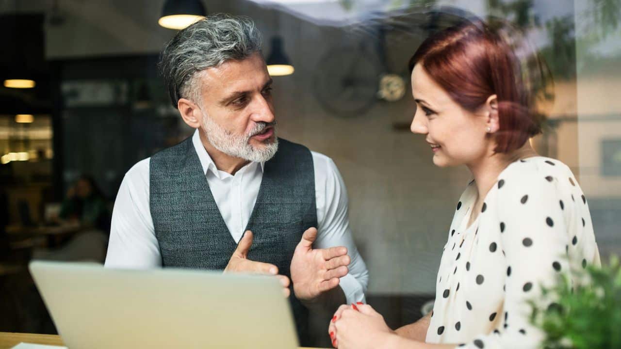 A grey-haired man in a vest talks animatedly to a red-haired woman at a table with a laptop.