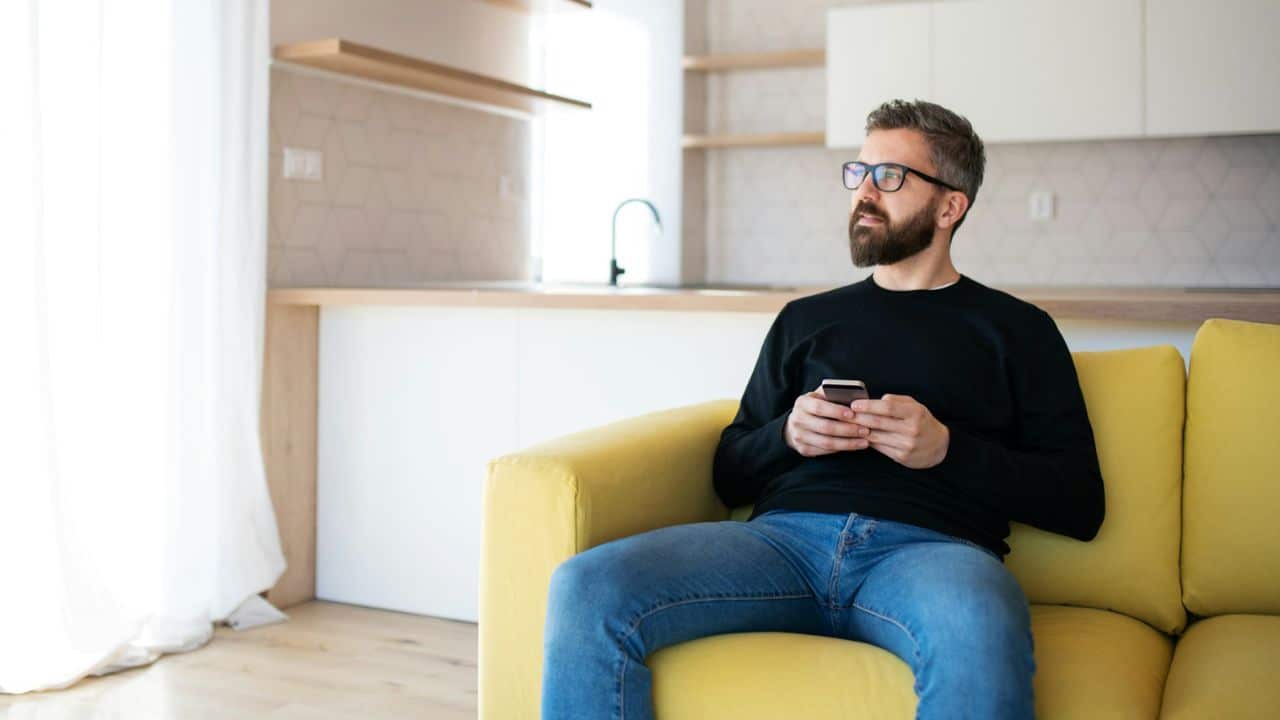 A bearded man in glasses and a black shirt sits on a yellow couch, holding a phone and looking away.
