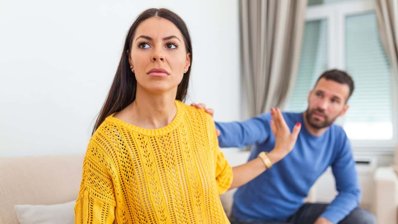 A woman in a yellow sweater looks away with a serious expression as a man in a blue shirt reaches out toward her.
