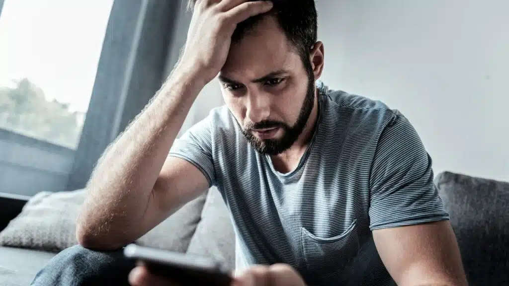 A bearded man in a striped t-shirt sits with his hand on his head, looking intently at a phone.