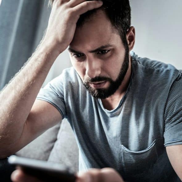A bearded man in a striped t-shirt sits with his hand on his head, looking intently at a phone.