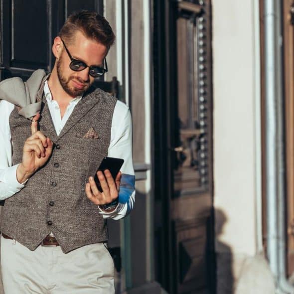 A man in a gray vest and sunglasses checking his smartphone on a sunny street.