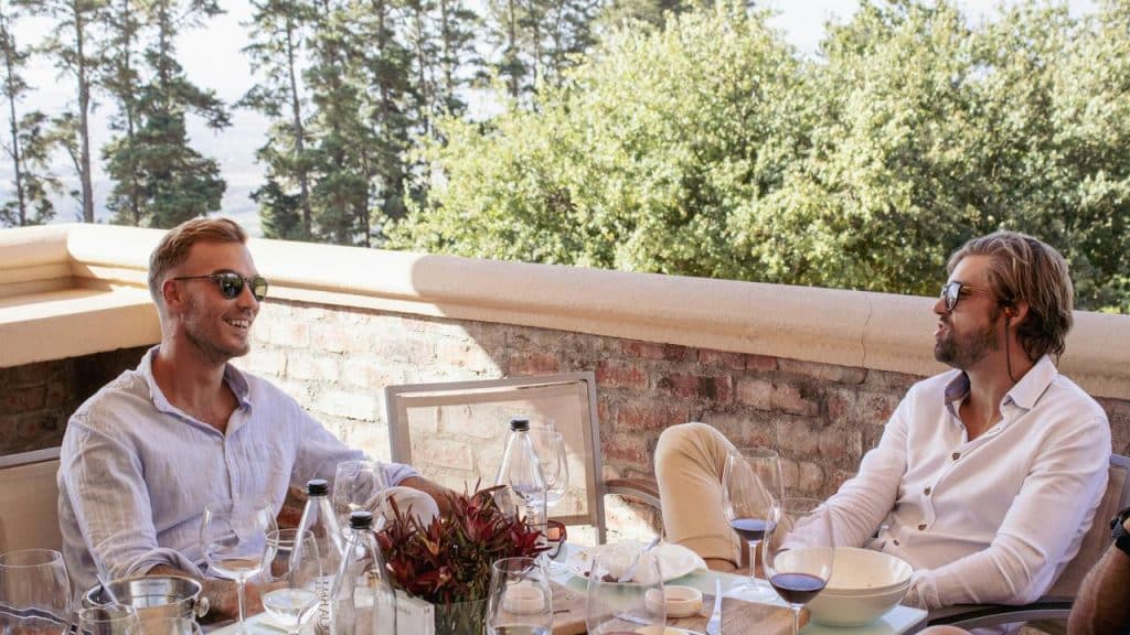 A pair of men wearing sunglasses chat over red wine at an outdoor terrace table.