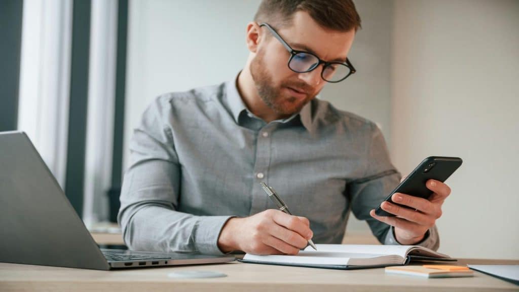 A man in glasses writes in a notebook while checking his phone at a desk.
