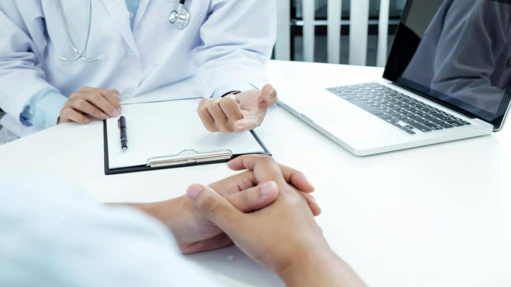 A doctor consulting with a patient at a desk with a laptop and clipboard.