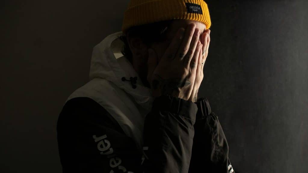 A person in a yellow beanie covers their face with both hands in dim light.