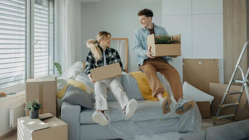 A smiling couple sits on a plastic-covered couch holding moving boxes.