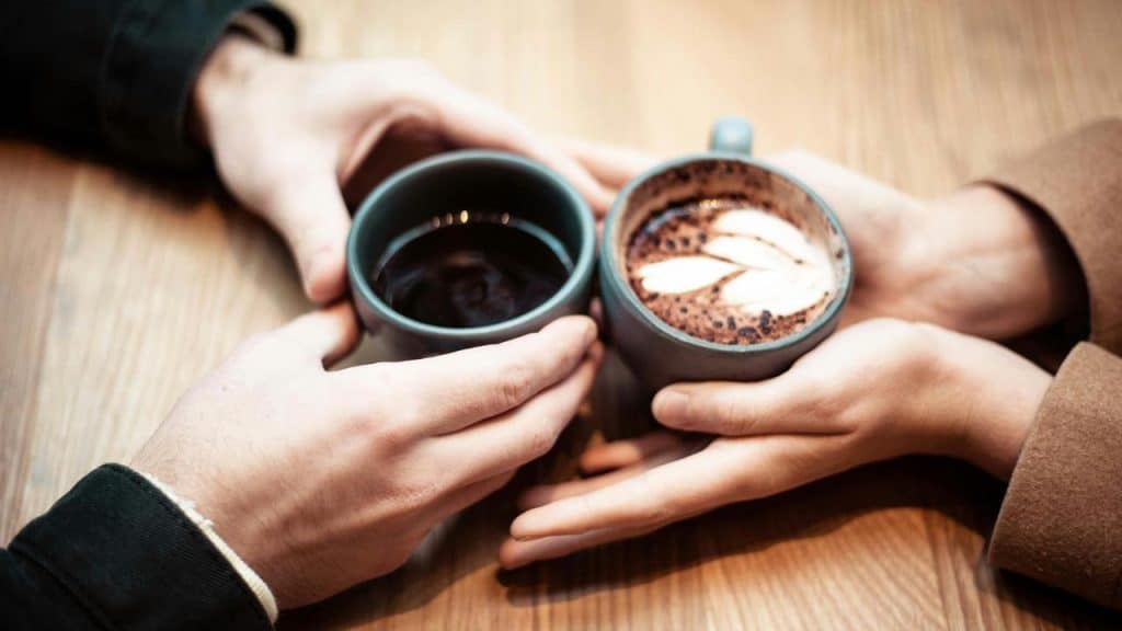 A pair of hands holding two coffee mugs, one black and one with latte art.