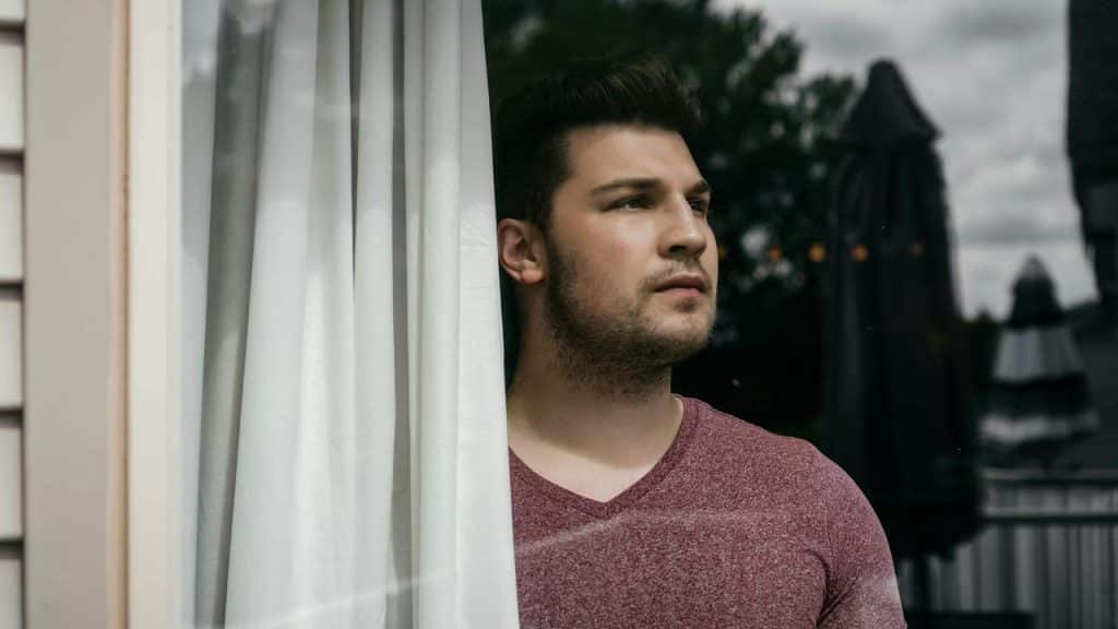 A man in a maroon shirt gazes out from behind a white curtain.