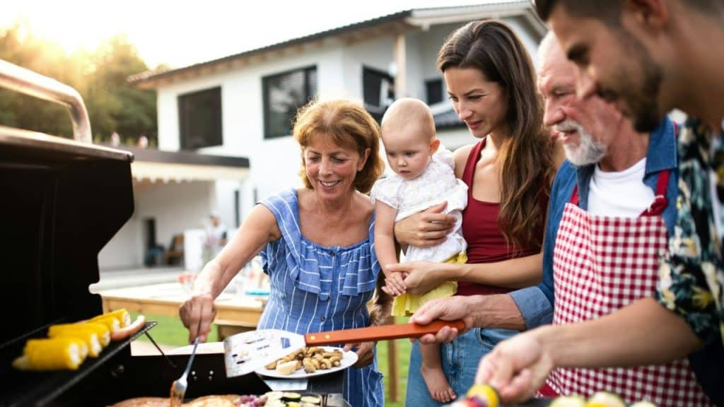 A multi-generational family is gathered around a grill outdoors.