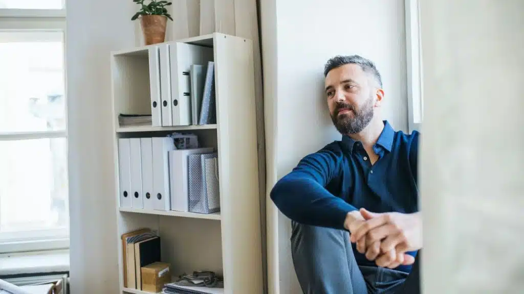 A pensive bearded man in a blue shirt sits on the floor next to a white bookshelf.