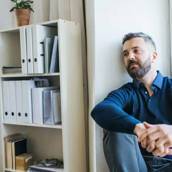 A pensive bearded man in a blue shirt sits on the floor next to a white bookshelf.