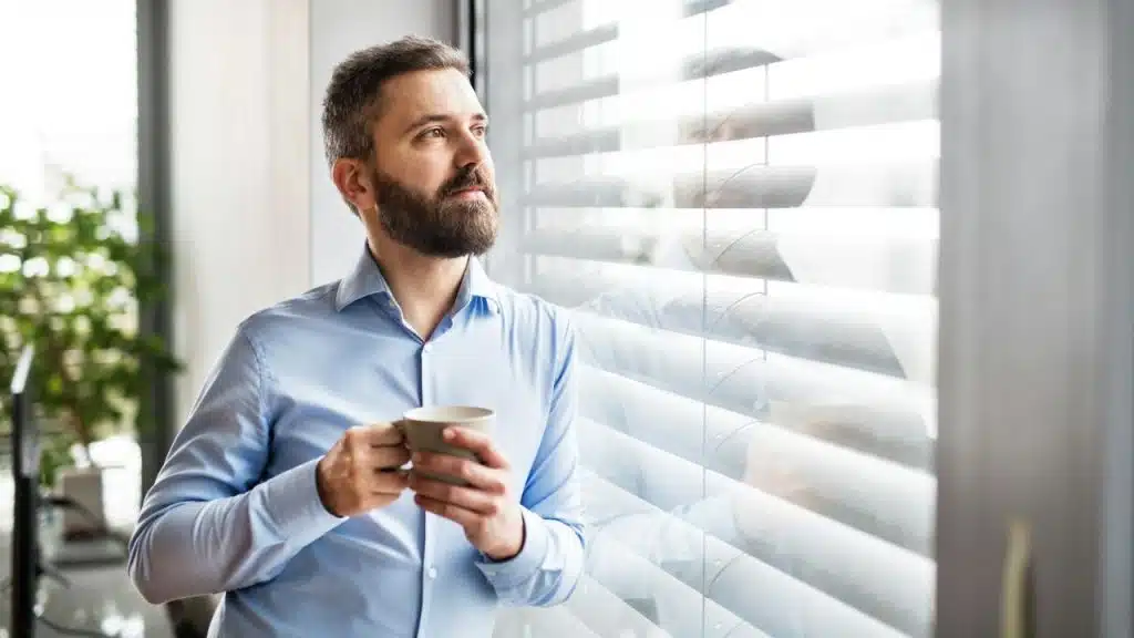 A bearded man in a blue shirt holds a mug and looks out a window with horizontal blinds.