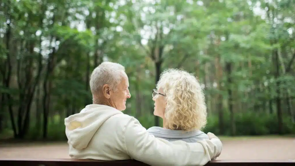 A smiling elderly couple sits on a bench in a park, looking at each other.