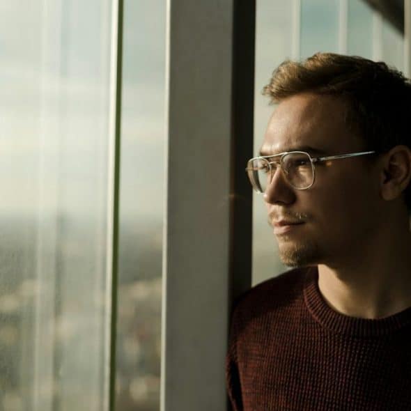 A man with short hair and glasses looks out a large window, bathed in sunlight.