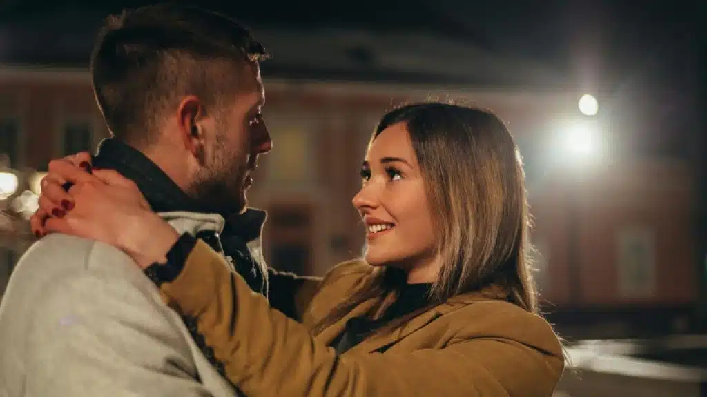 A man and a woman embrace outdoors at night, looking at each other with smiles.