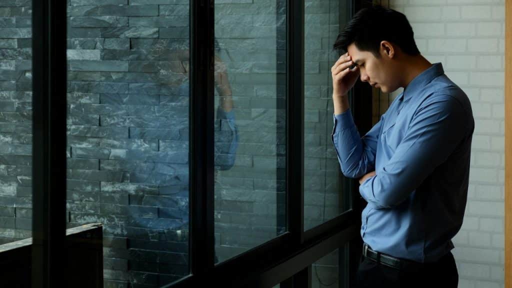 A man in a blue shirt stands by a window with his hand on his forehead, looking downcast.
