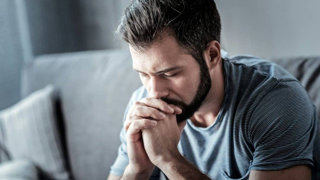 A bearded man with dark hair sits on a couch, looking down with his hands clasped.