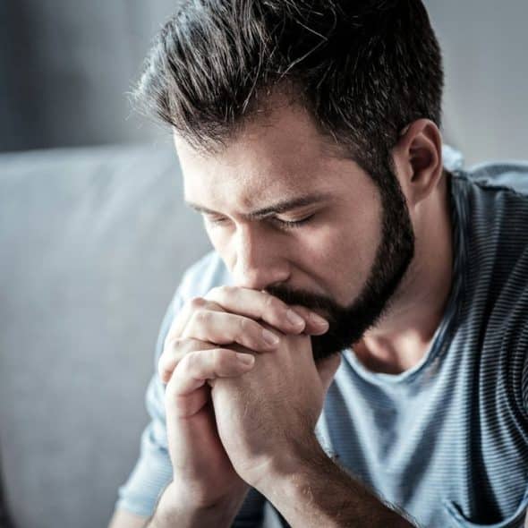 A bearded man with dark hair sits on a couch, looking down with his hands clasped.