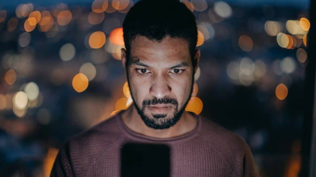 A serious man with a beard looks down at a phone screen, with blurred city lights in the background.