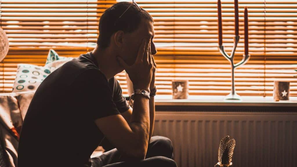 A man sits with his hands on his face, looking down, with a window and candles in the background.