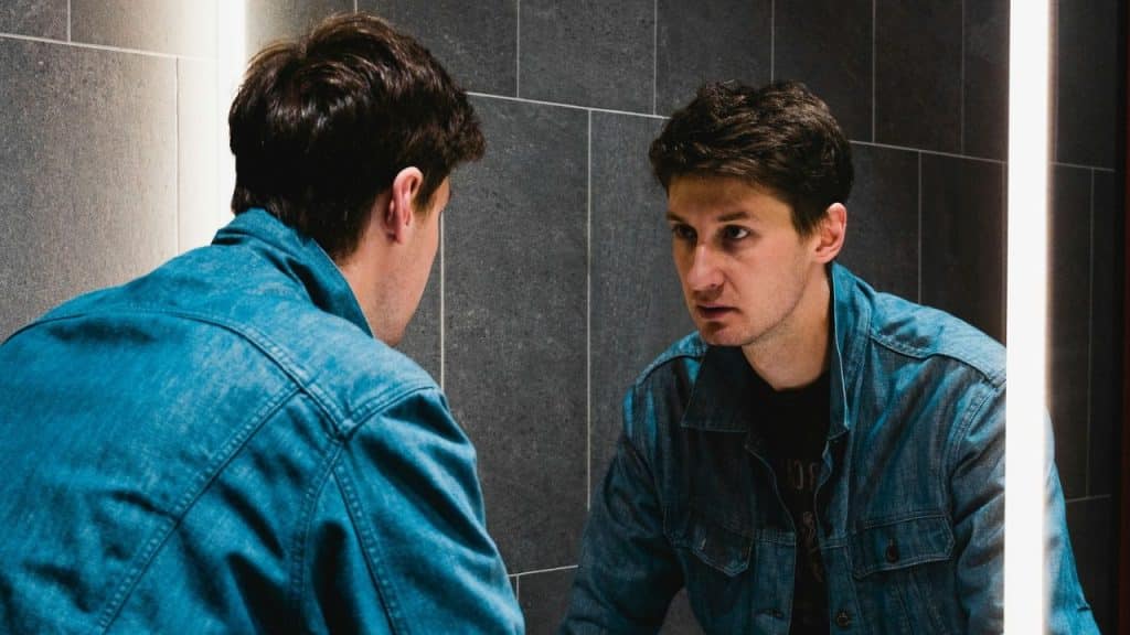 A man in a denim jacket looks at his reflection in a mirror, with a serious expression.