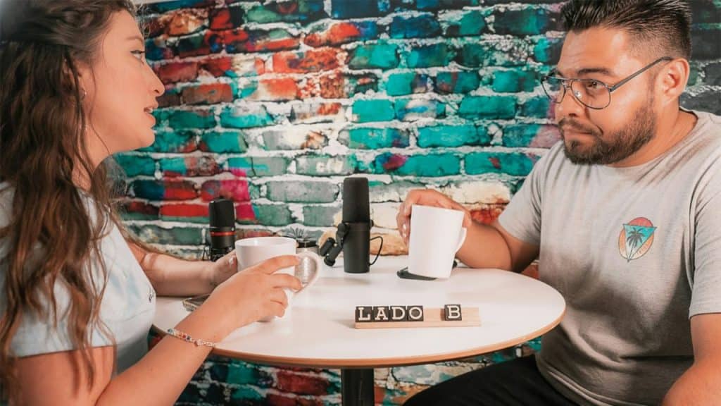 A woman and a man sit at a white table with microphones and mugs, facing each other in front of a brick wall.