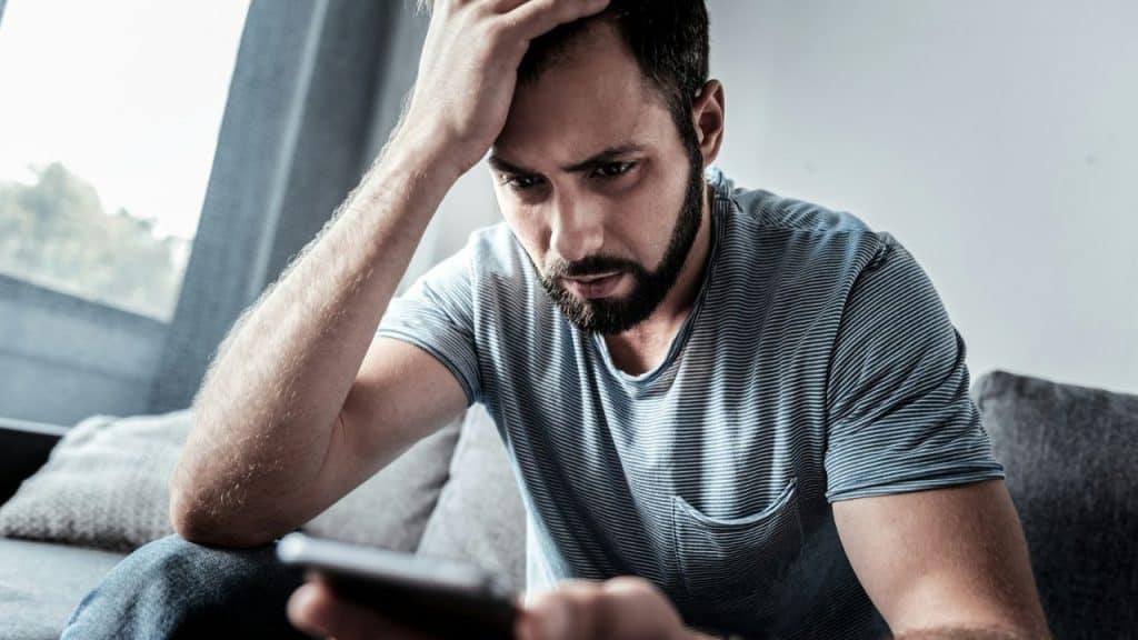 A bearded man with short dark hair sits on a couch, holding his head and looking intently at a phone.