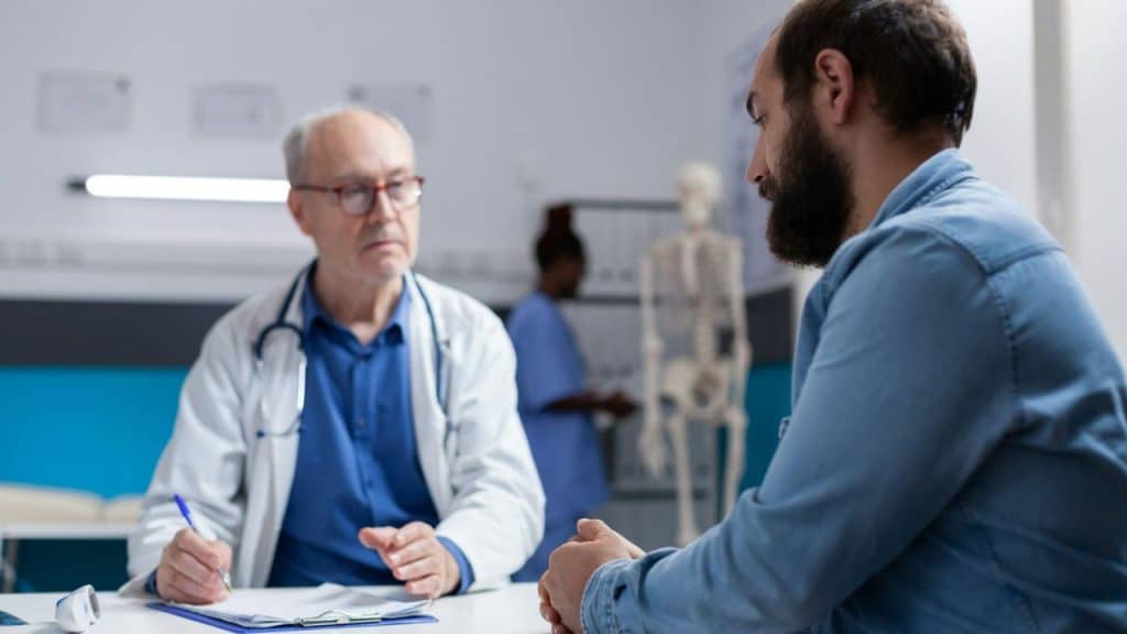 A male doctor in a white coat sits at a desk, writing on a clipboard, and looking at a bearded male patient sitting across from him.