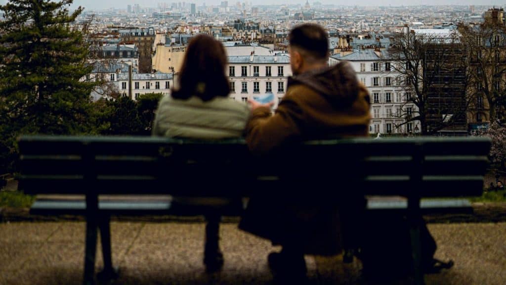 A man and a woman sit on a park bench looking out at a city skyline.