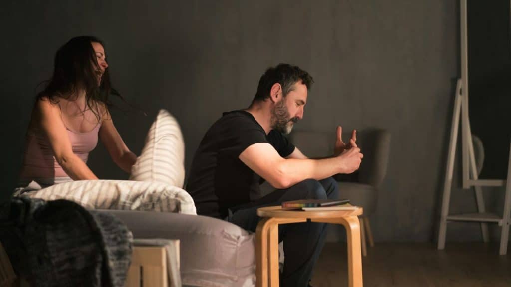 A woman sitting on a bed throws a pillow at a man with a beard who is sitting and looking down.