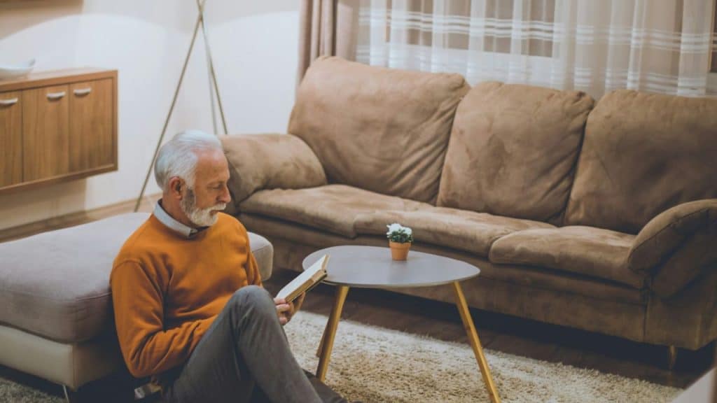 A white-haired man with a beard sits on the floor, reading a book next to a couch.