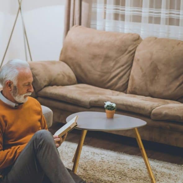 A white-haired man with a beard sits on the floor, reading a book next to a couch.