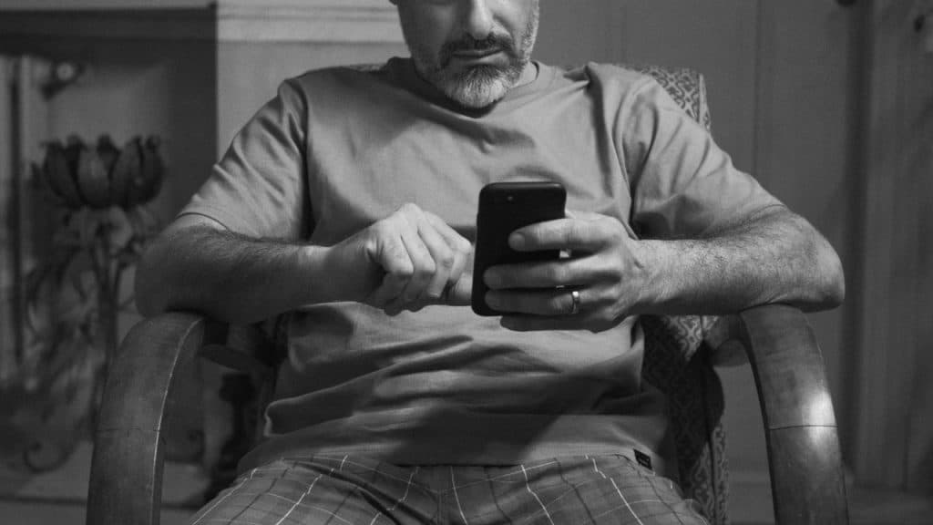 A bearded man sits in a chair, looking down and interacting with a smartphone.