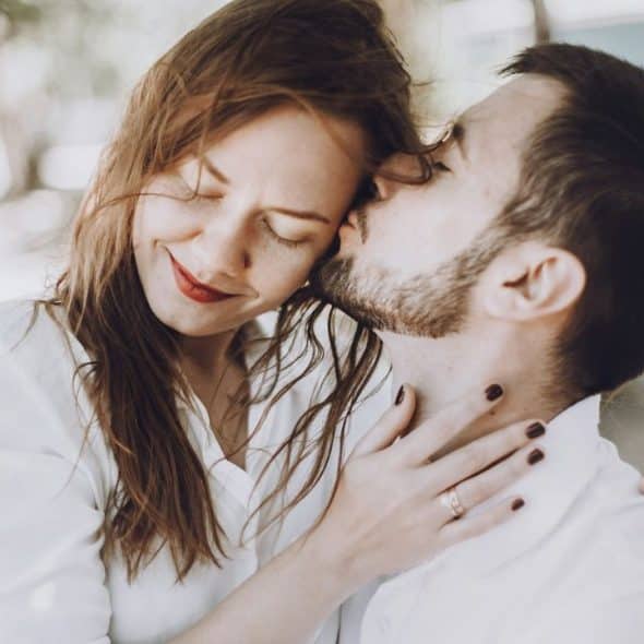 A man with a beard kisses a woman on the cheek, who has her eyes closed and a small smile, with her hand on his neck.